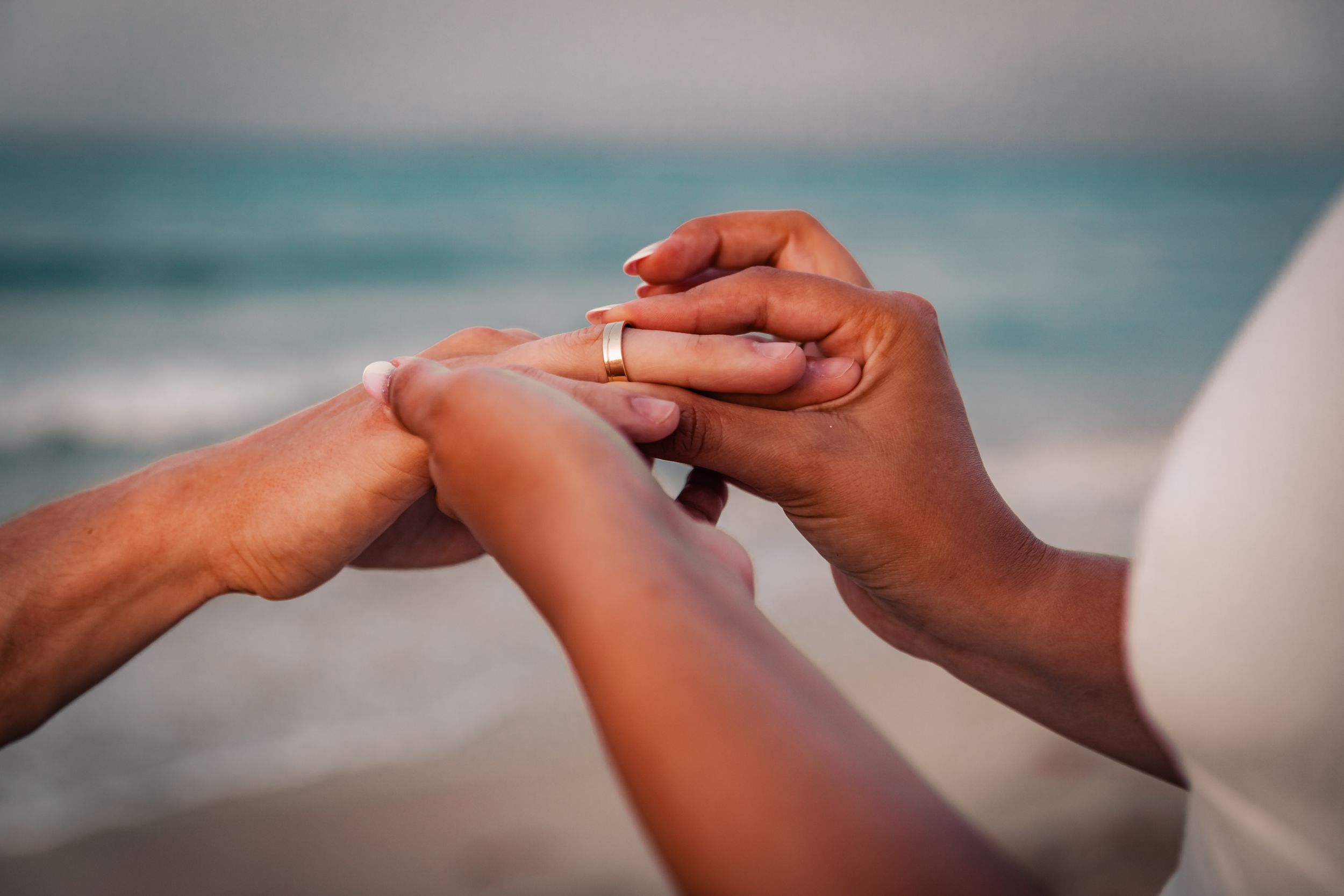 Close-up of a man placing an engagement ring on the woman's finger with blurred Mediteranean sea behind.