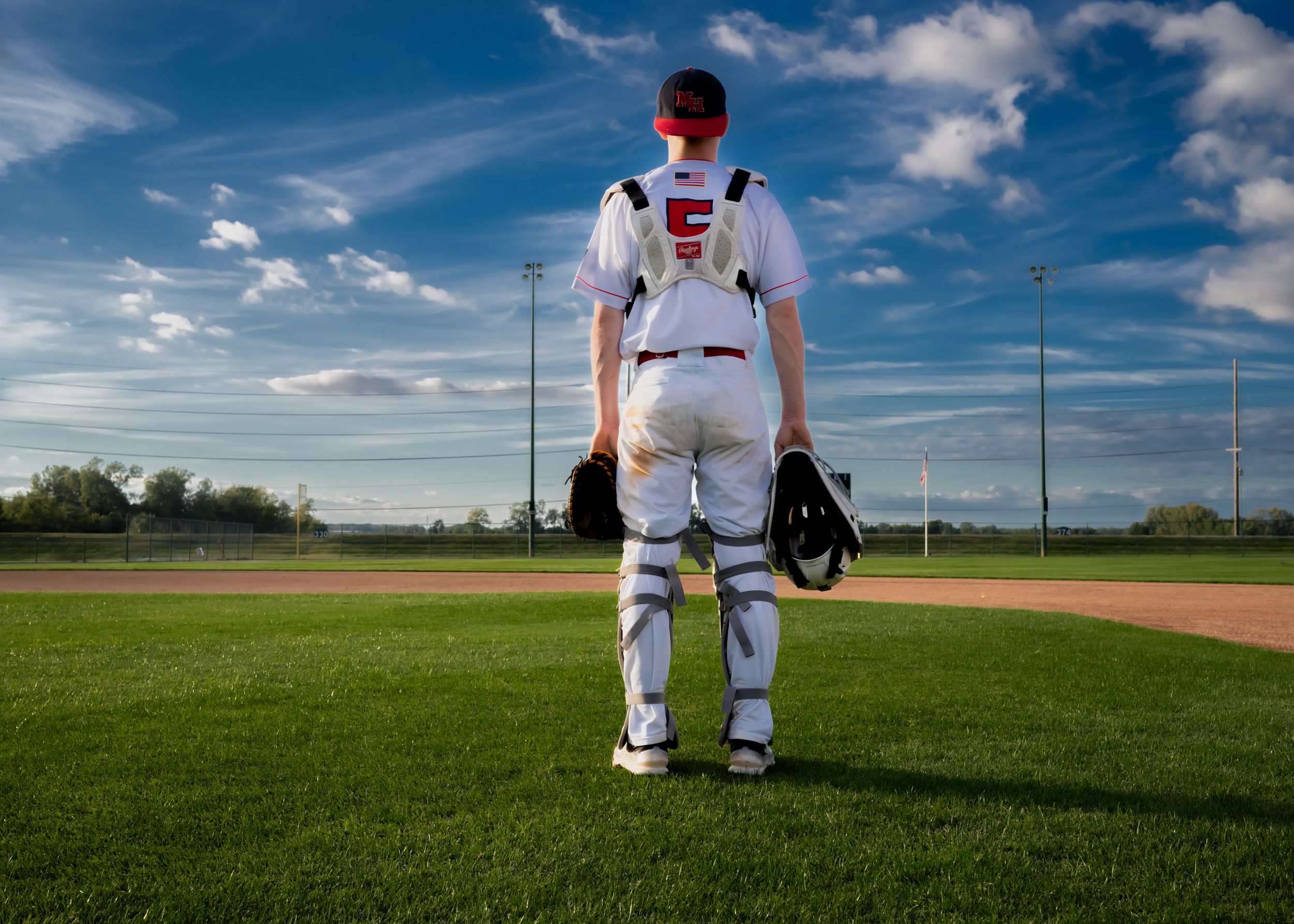 Baseball player in white uniform and gear stands on baseball field with blue sky background.