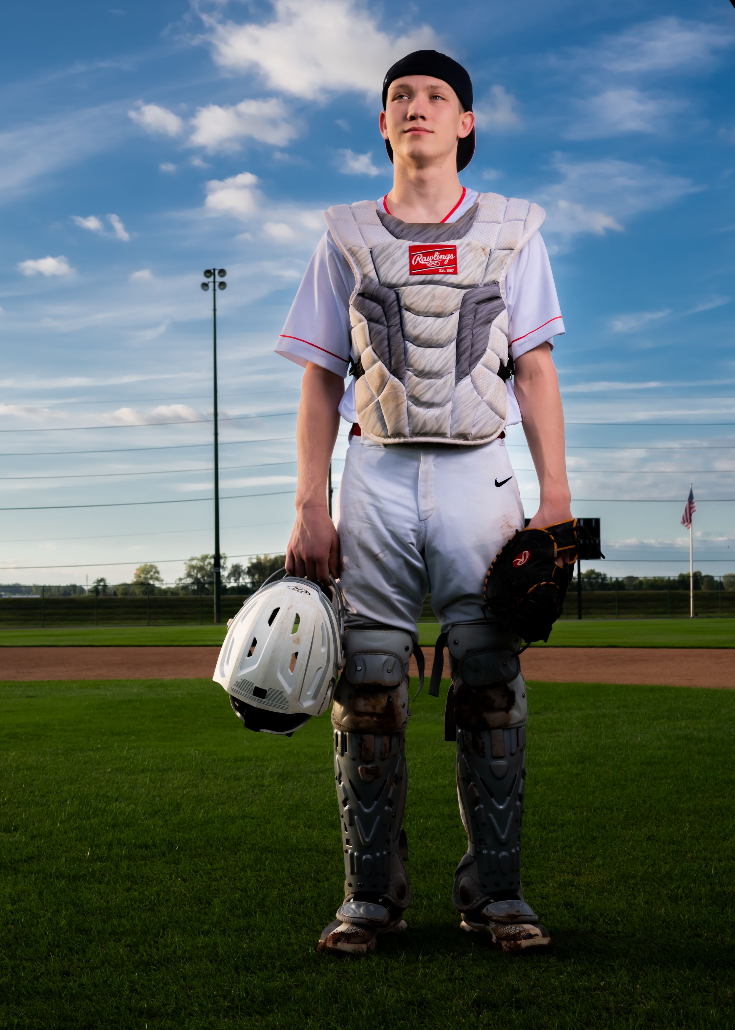 Baseball catcher in full white uniform and protective gear stands on field under blue sky with clouds.