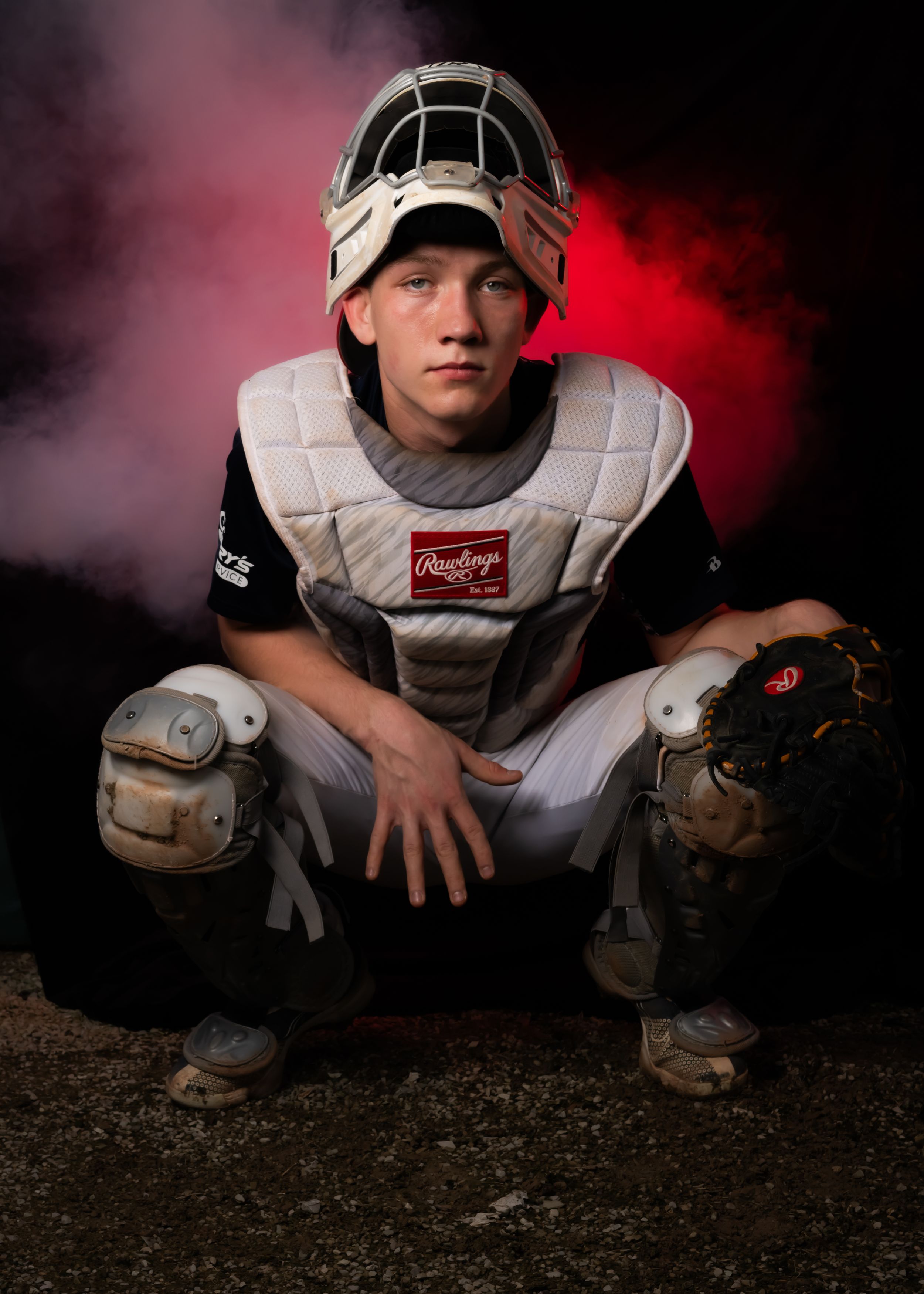 Baseball catcher in protective gear poses with dramatic red smoke effect backdrop.