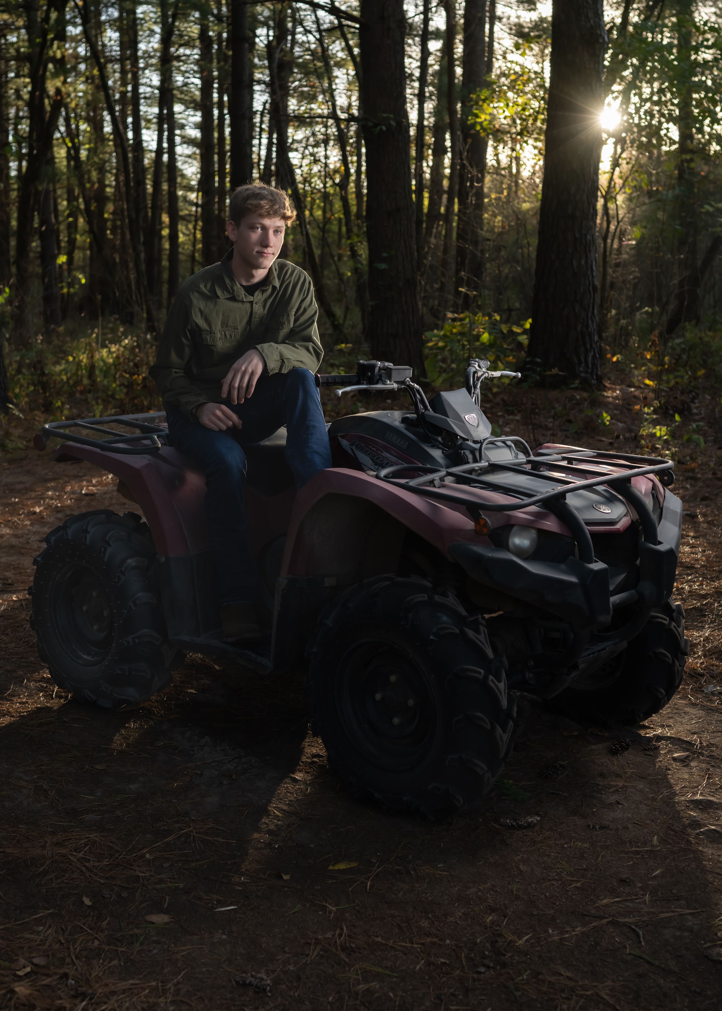 Person sits on red ATV on forest trail at sunset.