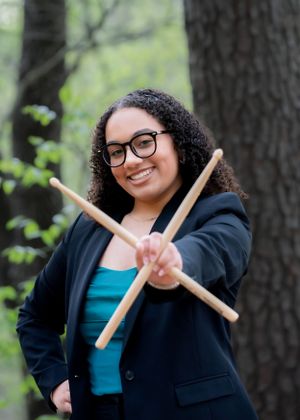 Musician in black blazer holding drumsticks in wooded outdoor setting.