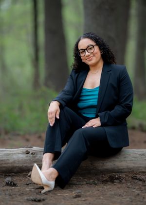 Professional woman in business attire sitting casually on a log in a wooded setting.