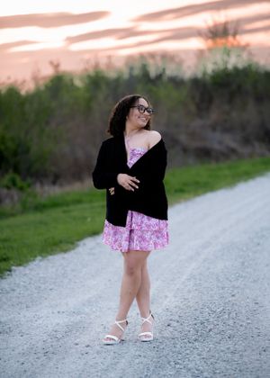 Person in pink dress and black cardigan posing on rural road at sunset.