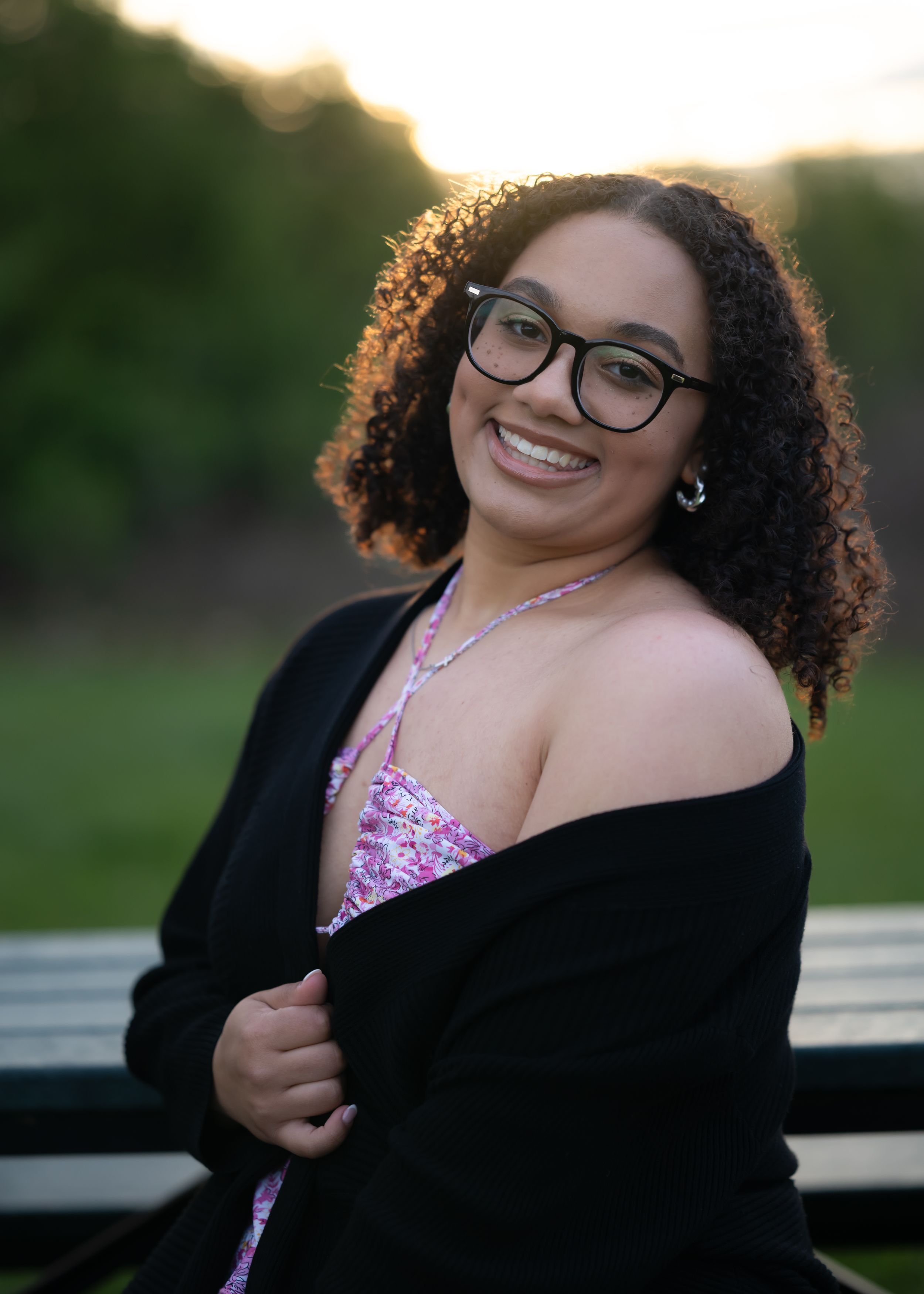 Portrait in black cardigan and pink top against sunset with natural backlit curls.