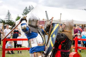 Medieval knights in armor clash with swords and shields during a historical reenactment battle at an outdoor festival.