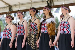 Performers wearing matching geometric patterned tops and black skirts stand in formation at an outdoor cultural festival.
