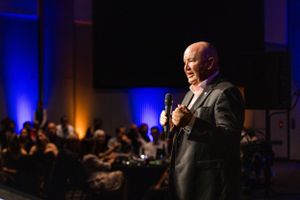 A speaker in a dark suit stands on stage with a microphone against dramatic blue lighting at a formal event.