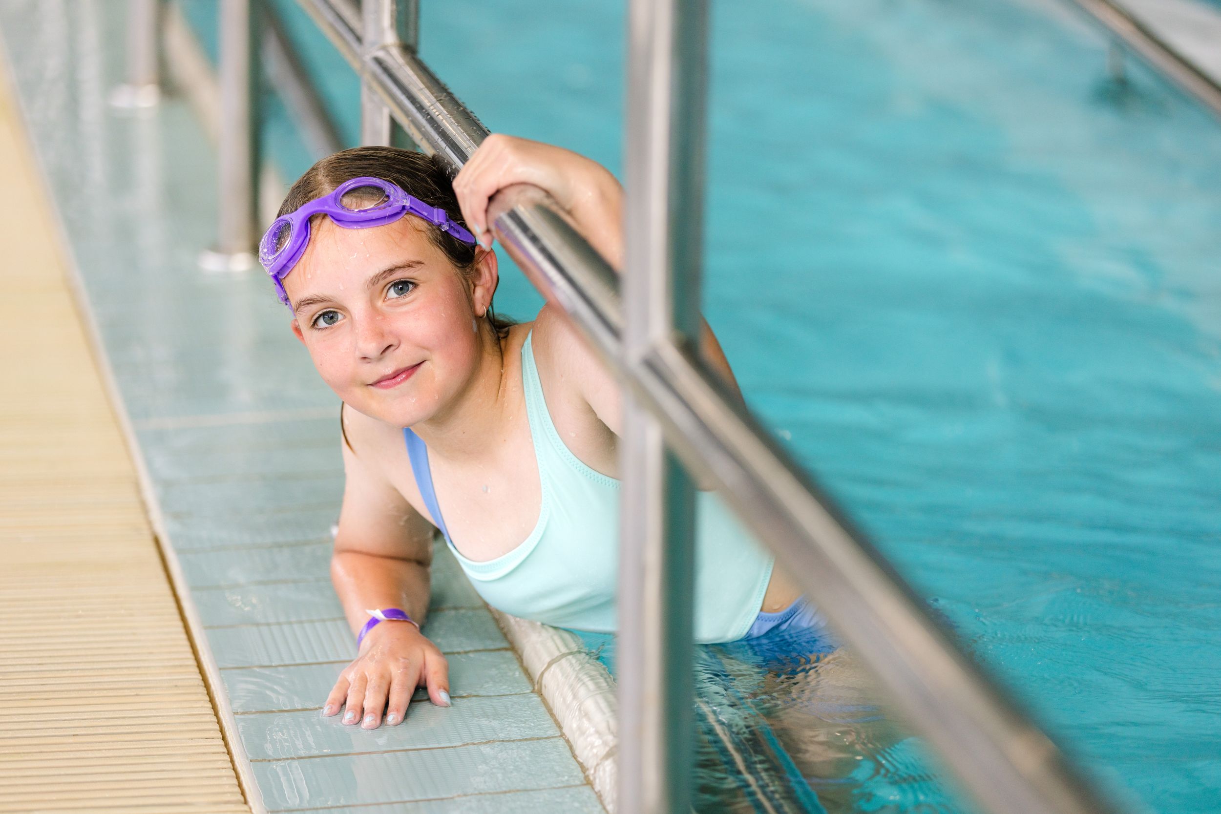 Swimmer in light blue swimsuit with purple headband holds onto pool edge during swimming lesson.