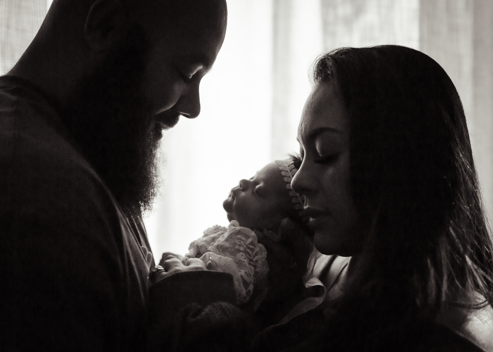 A silhouetted couple gazes lovingly at their newborn baby in a tender black and white portrait.