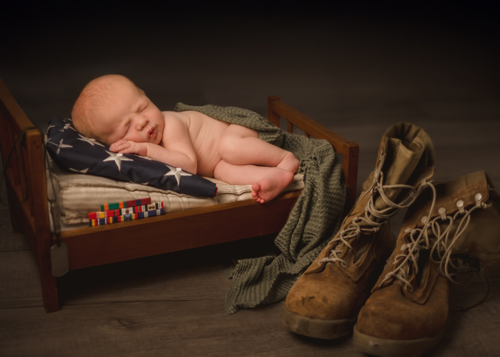 A sleeping newborn baby lies on a wooden surface next to military combat boots and an olive green blanket.