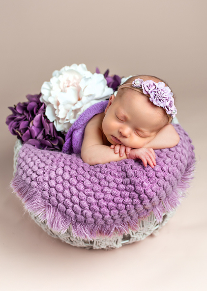 Sleeping newborn wearing purple headband on textured knit blanket with purple flowers in basket.