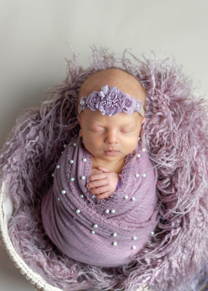 Swaddled newborn on purple fuzzy blanket wearing pearl-adorned headband.