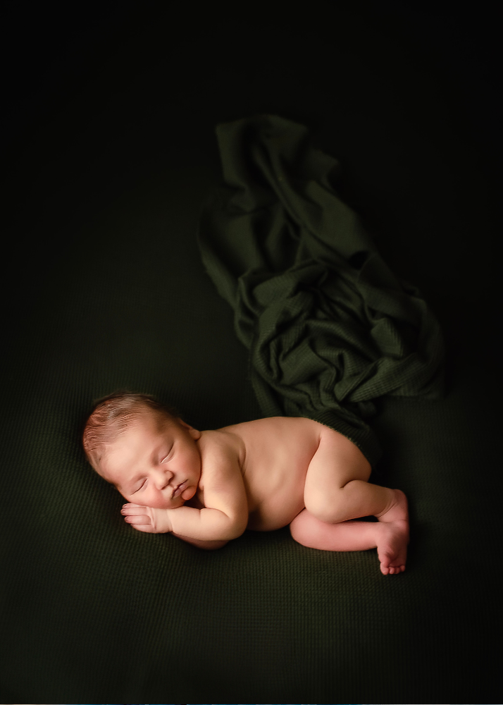 Peaceful newborn sleeping on dark green fabric in artistic studio portrait.