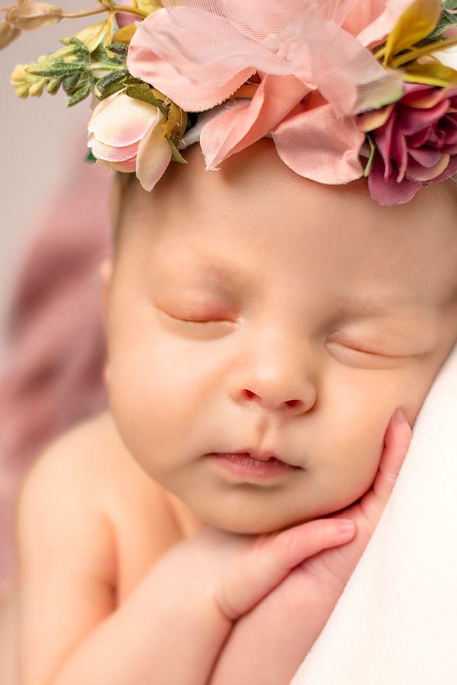 Peaceful sleeping baby wearing delicate pink and mauve floral crown against soft pink background.