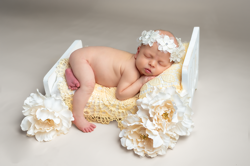 Sleeping newborn lying in white basket with cream colored flowers and delicate lace headband on beige background.