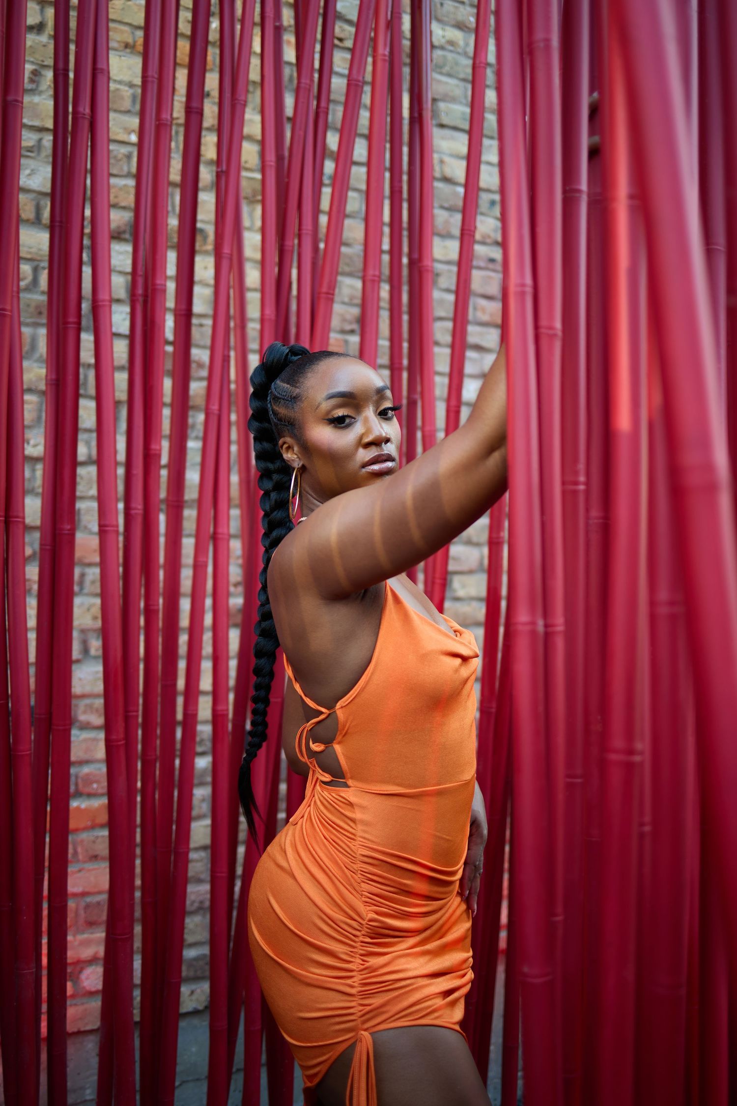 Woman in orange dress posing dramatically against red bamboo backdrop.