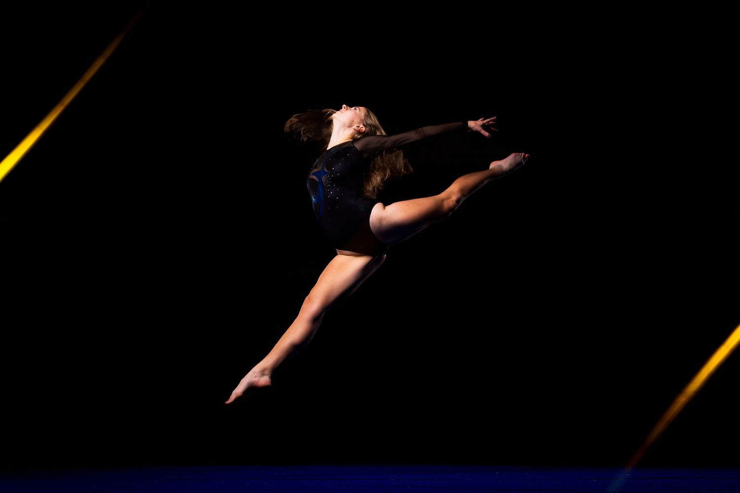 Graceful dancer in black leotard performs dramatic split leap against dark background with diagonal yellow light beams.
