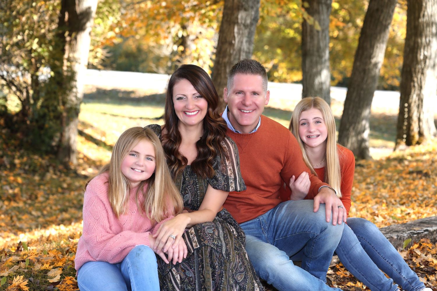 Family of four sitting together outdoors during autumn with trees in background during fall photo session.