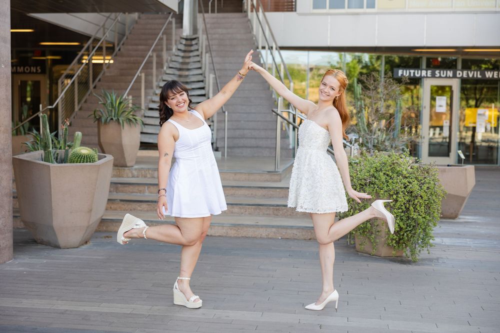 Two friends in white dresses dancing and twirling on urban steps.