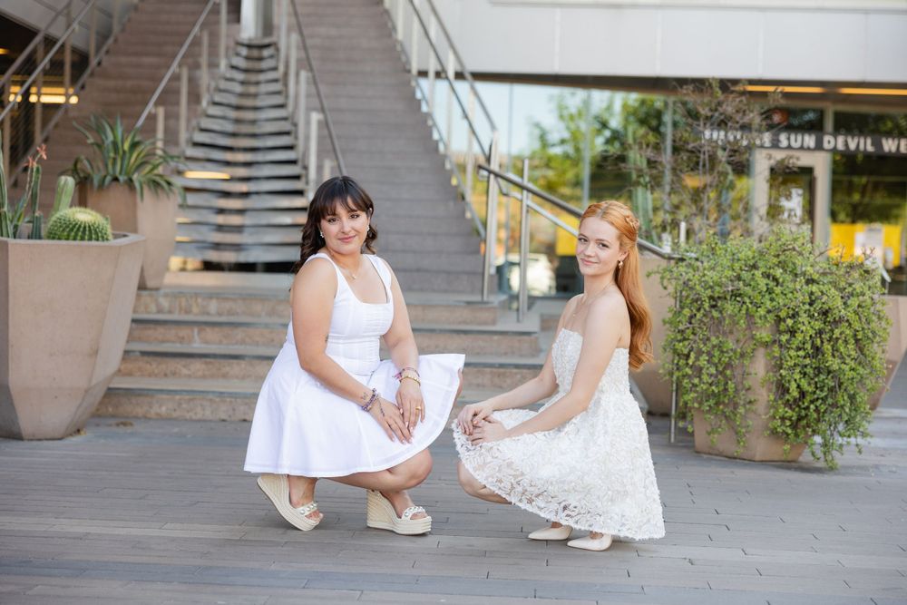 Two friends in white dresses posing together on urban steps.