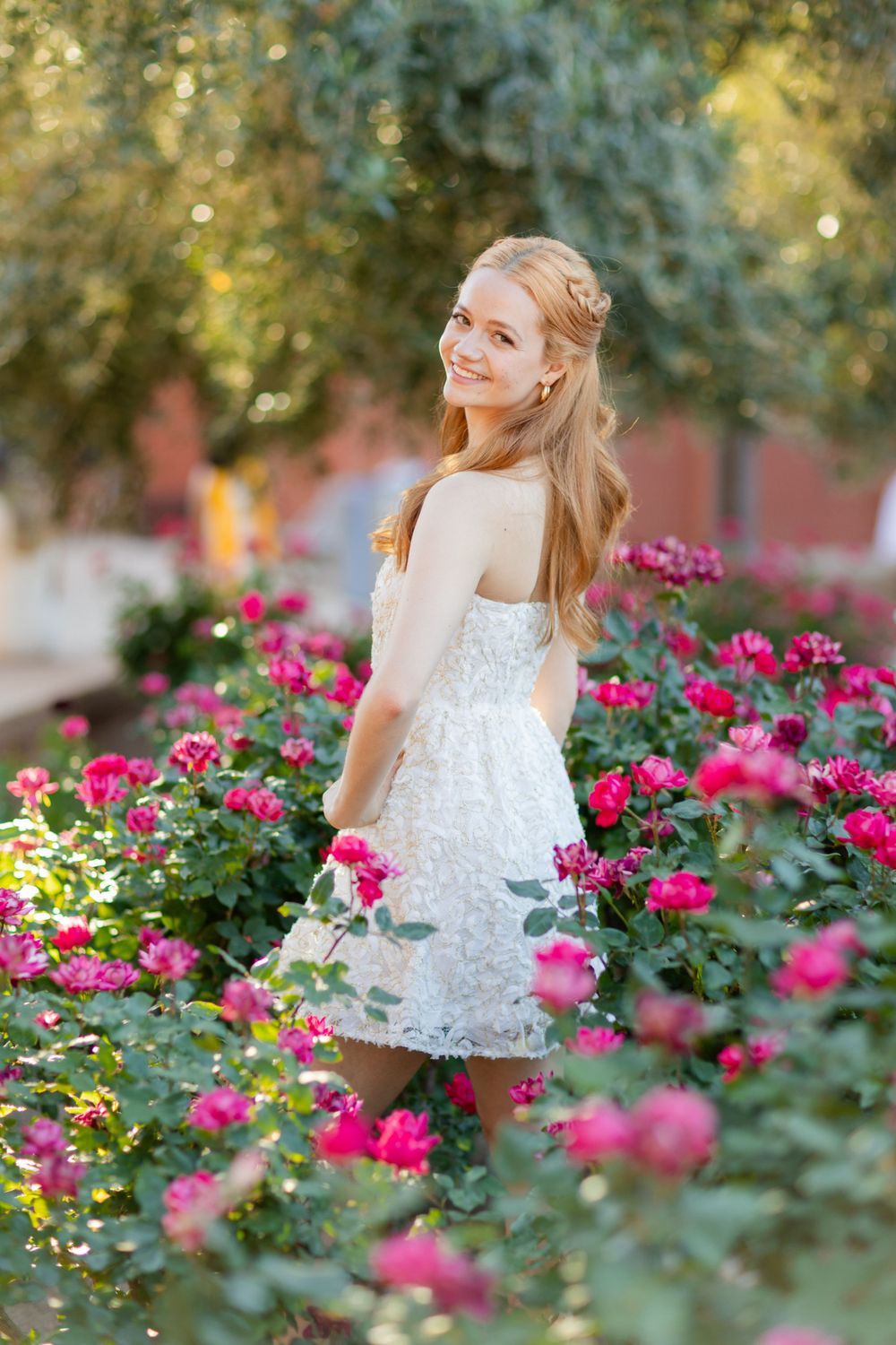 Person in white dress smiles while posing among blooming pink rose bushes.