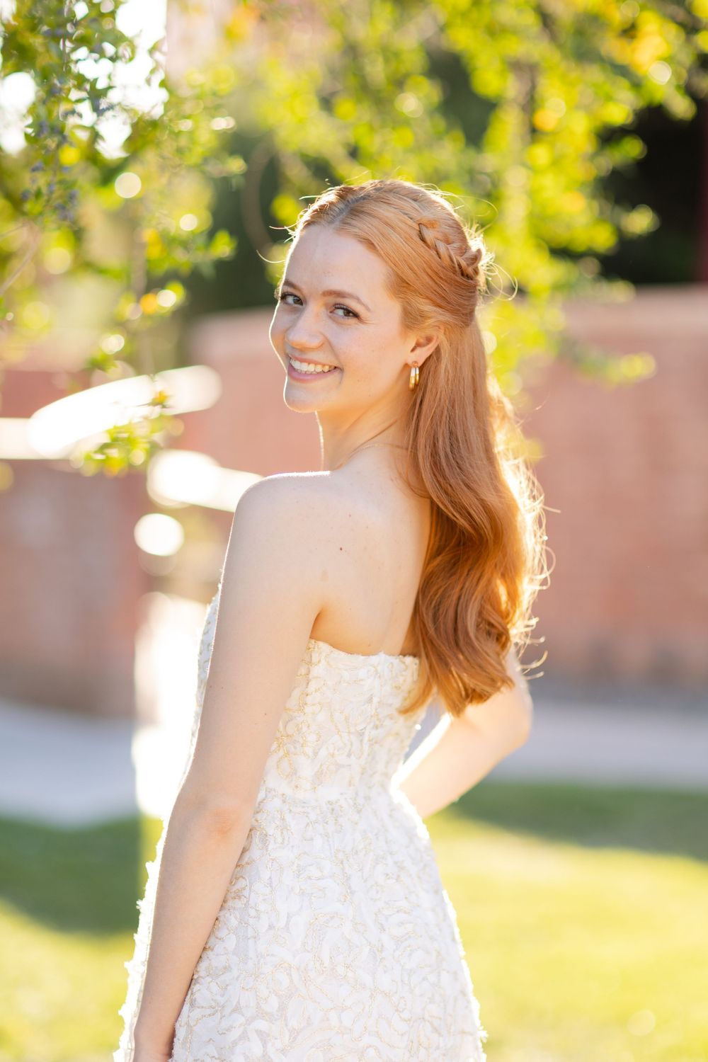 Elegant white lace dress captured in golden sunlight with brick wall and foliage backdrop.