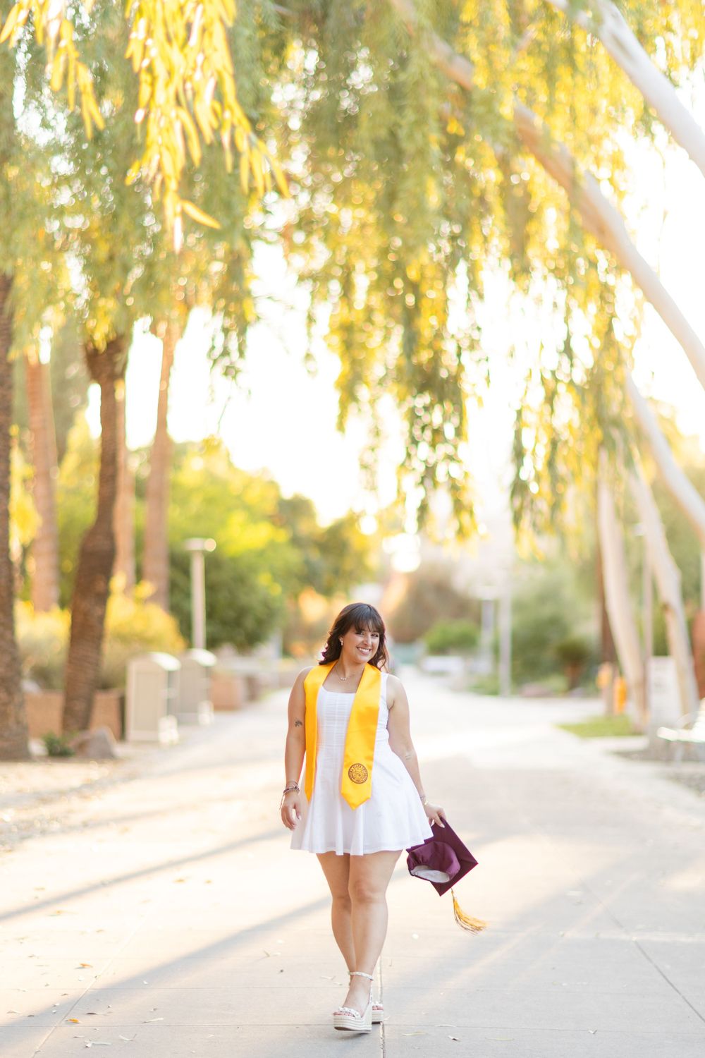 Graduate strolling down campus pathway under willow trees in white dress and gold stole.