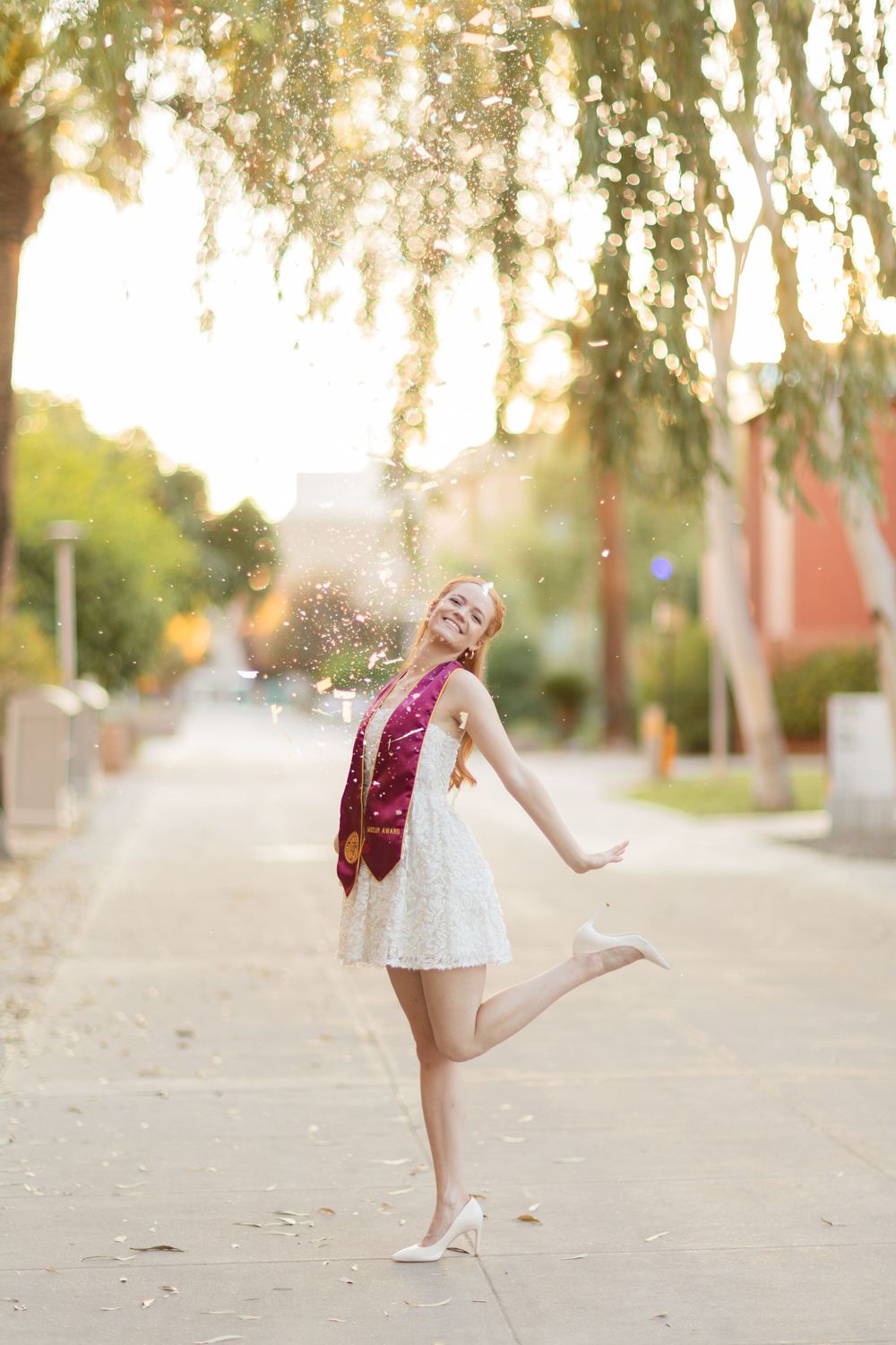 Graduate in white dress and maroon stole dances joyfully under willow trees.