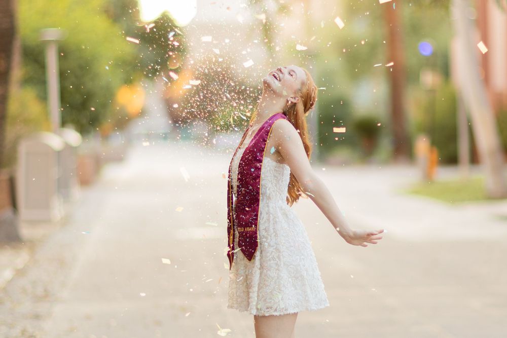 Graduate celebrating under trees with glitter and maroon academic regalia.