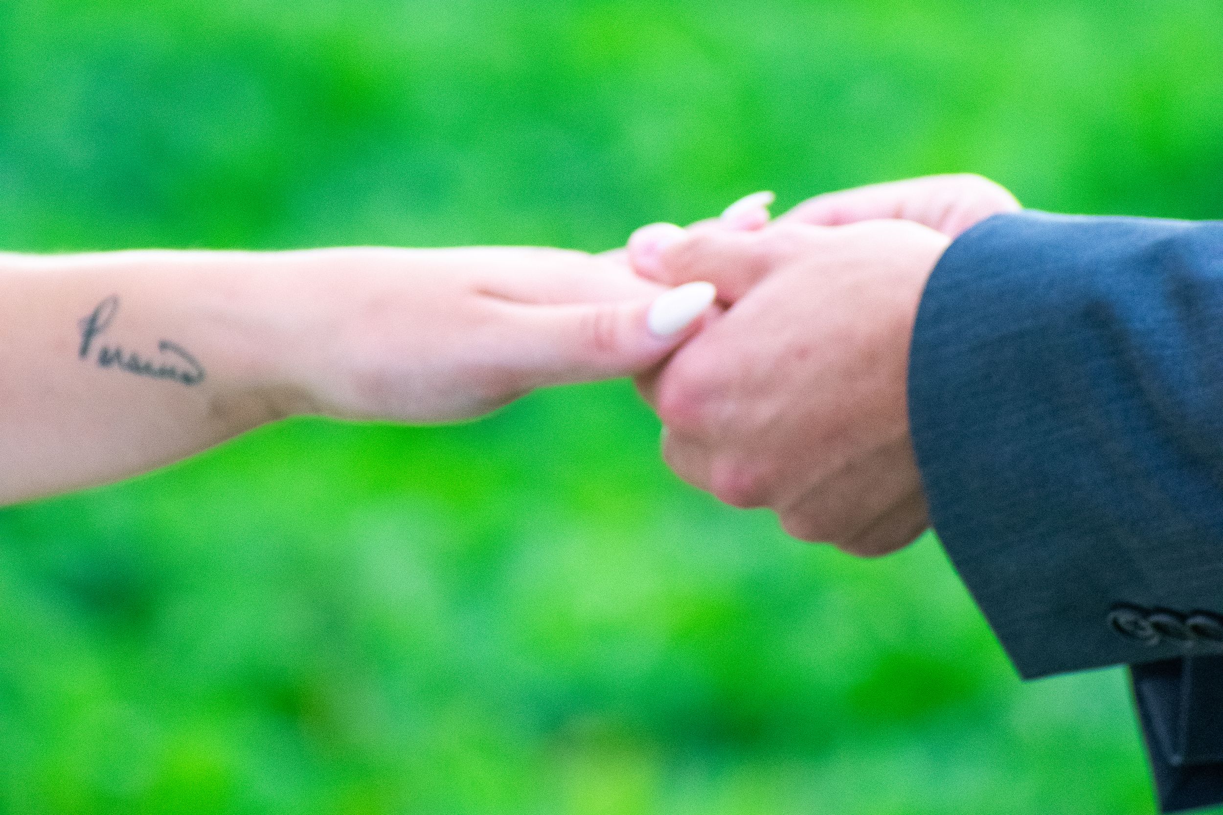Close-up of hands with visible tattoo during wedding ring exchange on green background.