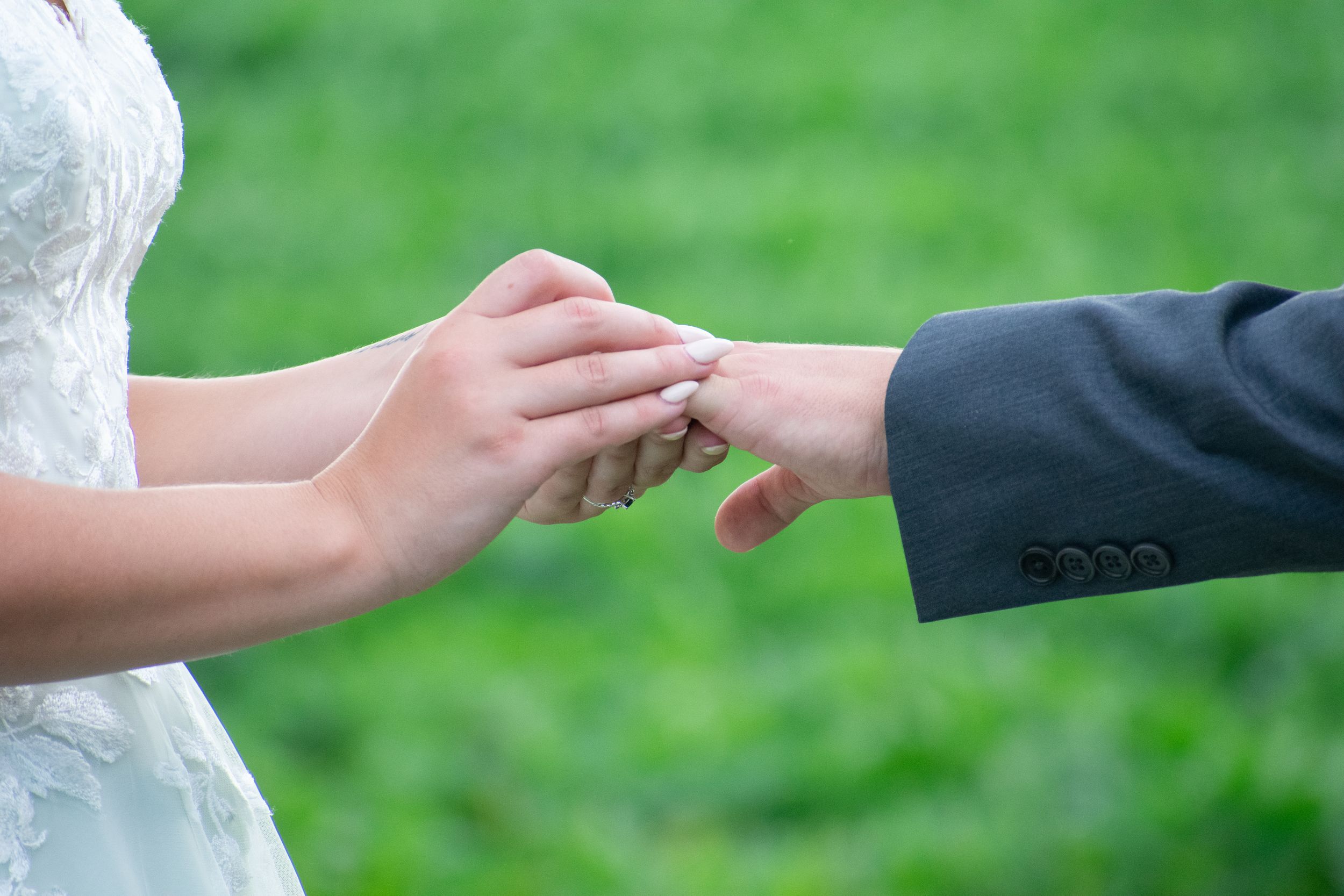 Close-up of hands exchanging wedding rings against green outdoor background.