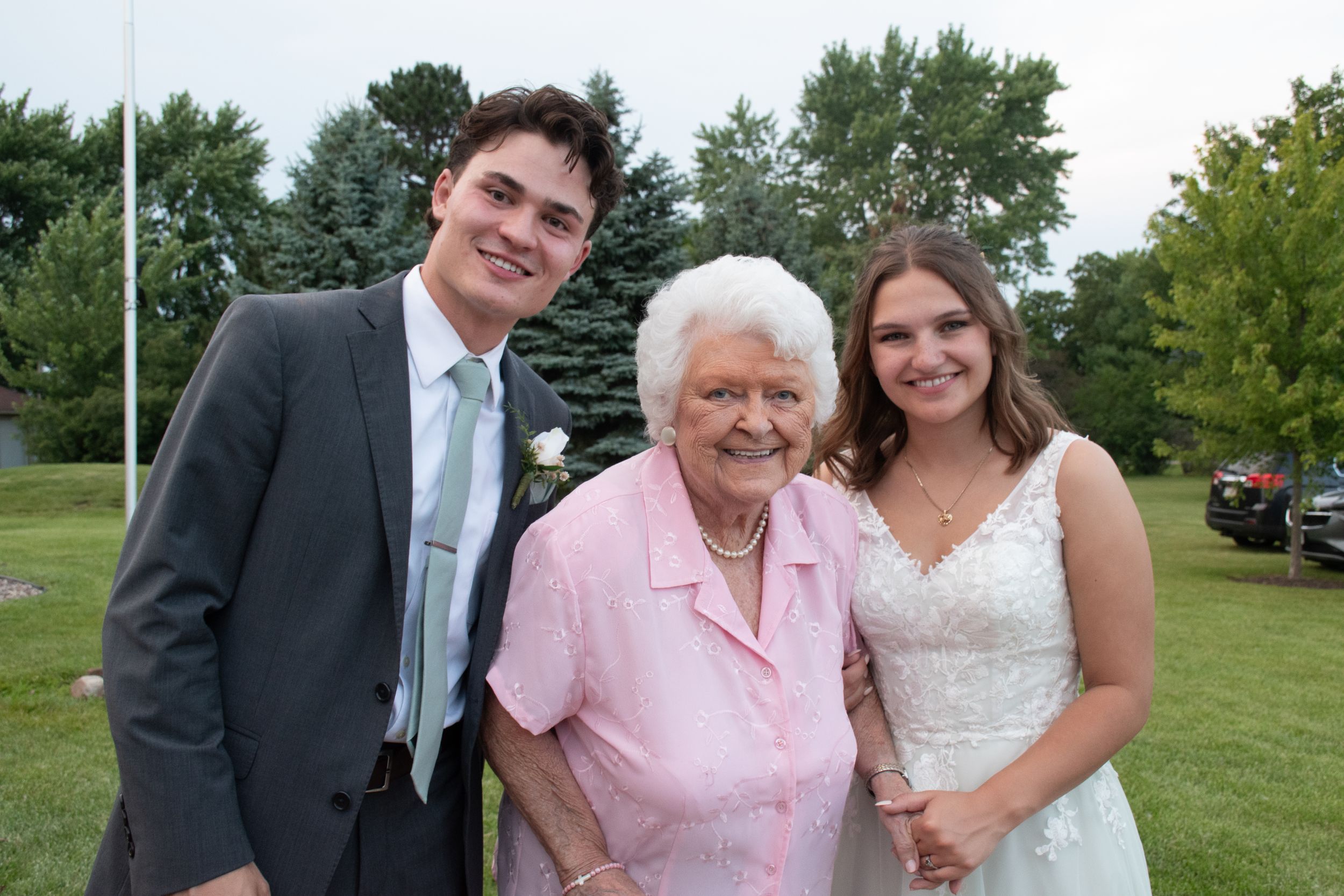 A wedding couple poses with an elderly relative in formal attire outdoors.