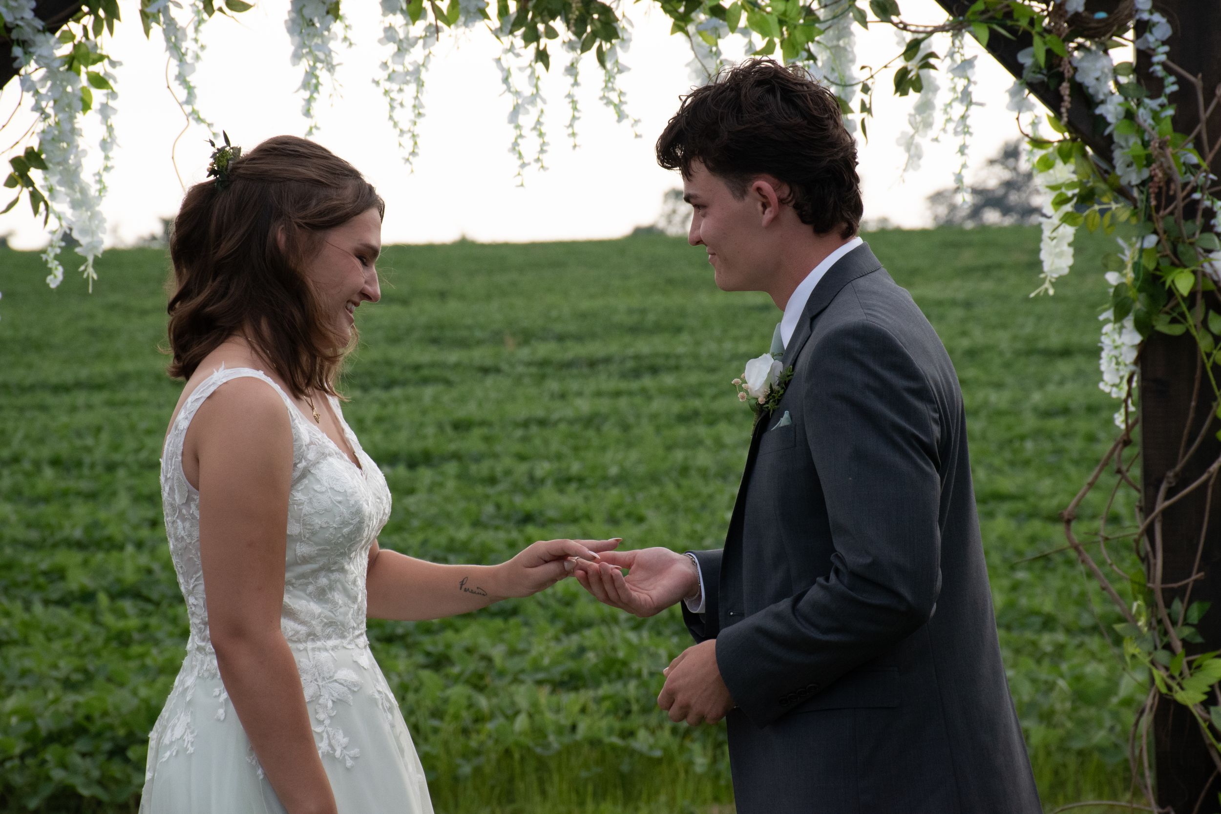 A couple exchanges rings under a floral arch during an outdoor wedding ceremony.