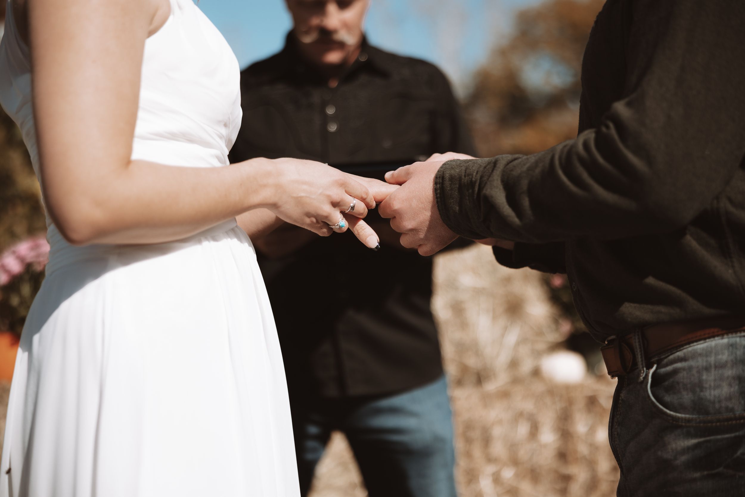 Close-up of hands exchanging rings during an outdoor wedding ceremony with person wearing white dress.