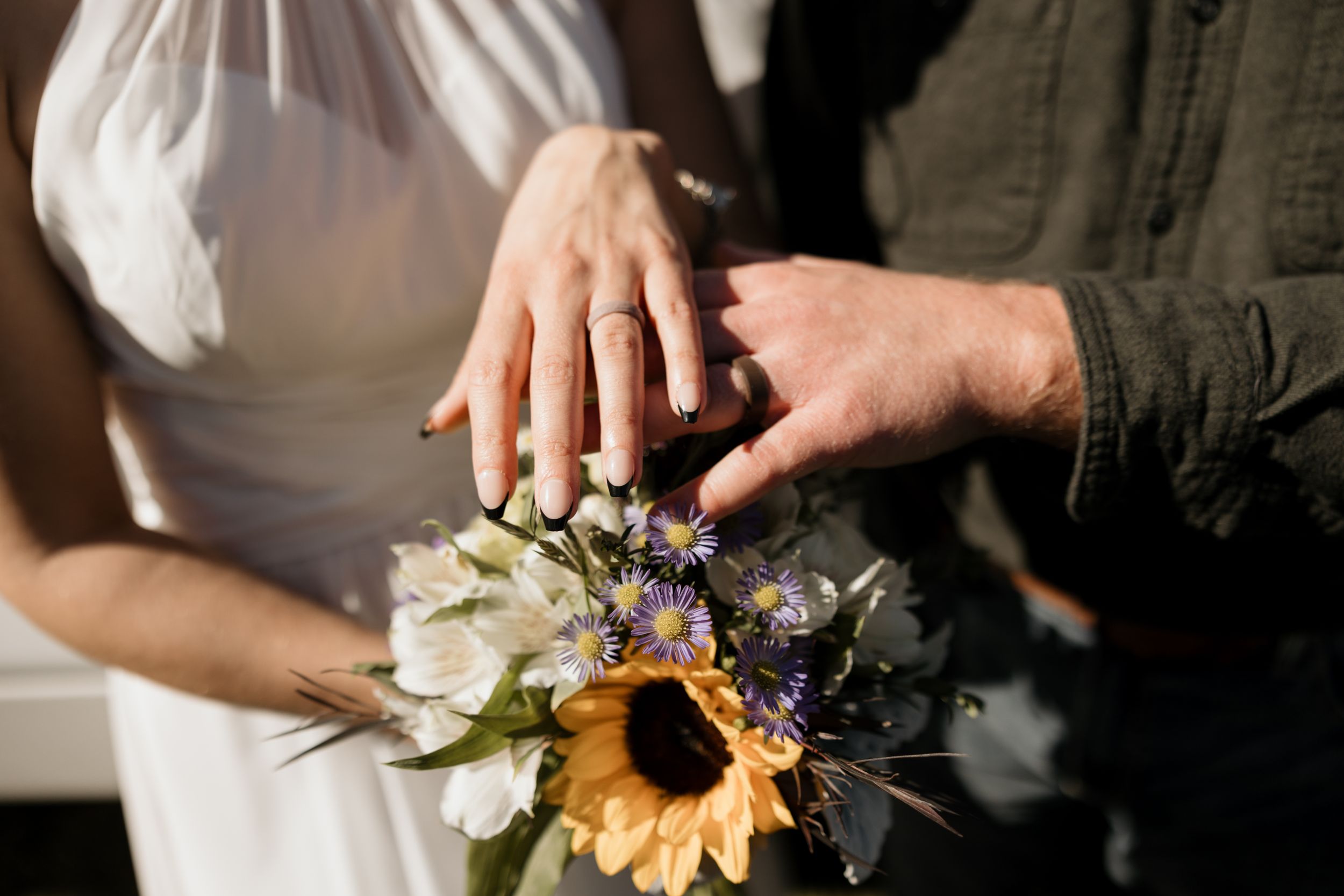 Two hands rest together over a rustic bouquet featuring a sunflower and wildflowers in soft natural lighting.
