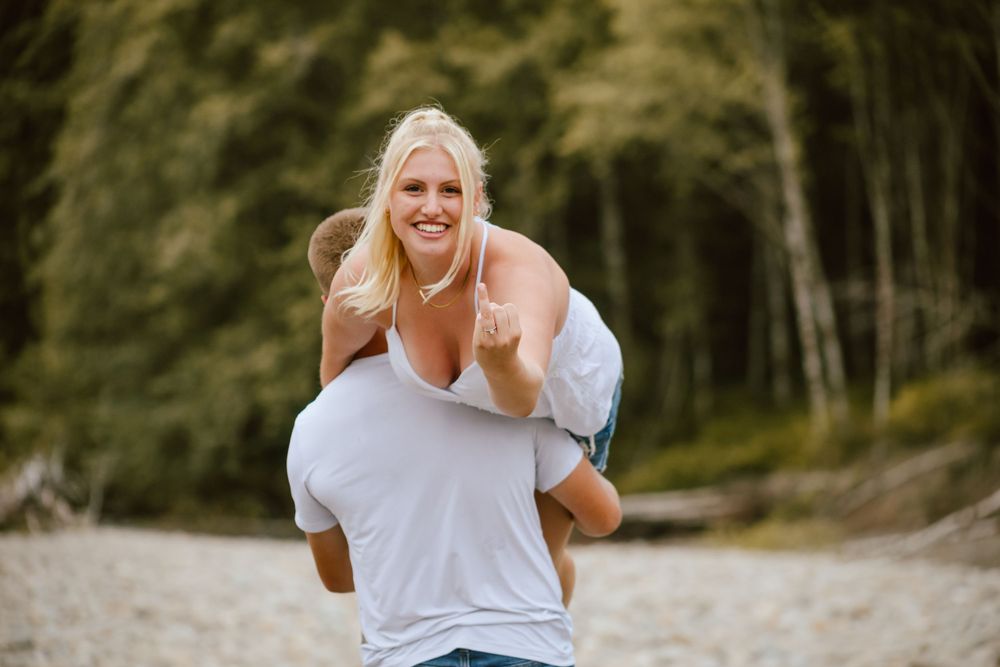 Playful piggyback ride showcasing her engagement ring captured on a river bed with forest background.