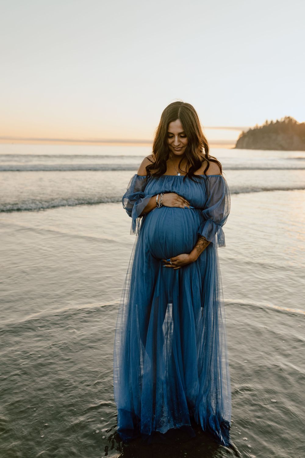 Silhouette of pregnant woman in flowing blue dress standing at sunset beach.