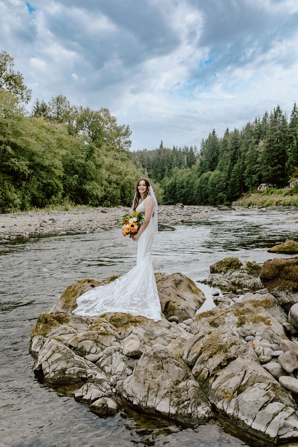 Person in white wedding dress holding bouquet while standing on rocky riverbank.