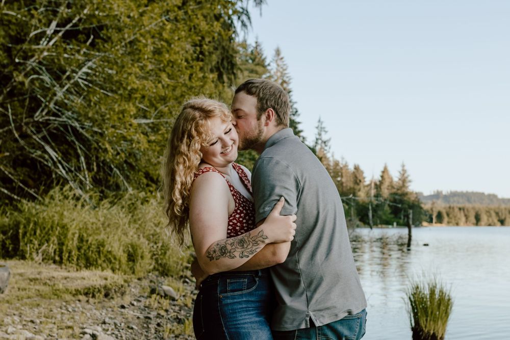 Romantic couple embracing by scenic lakeside with forest in background.