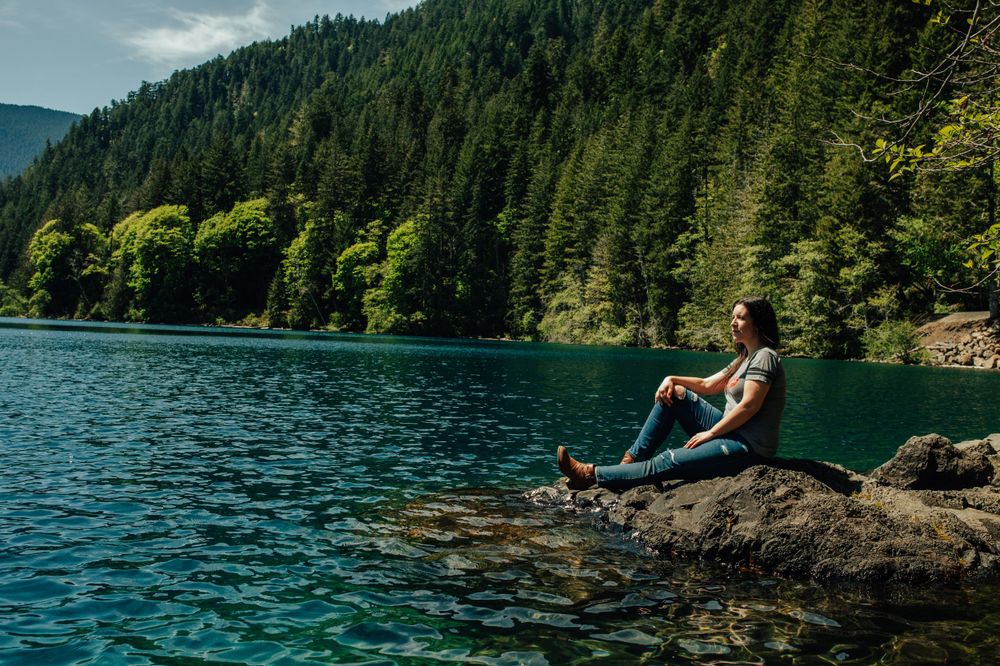 Person relaxing on rock overlooking pristine turquoise mountain lake surrounded by evergreens.