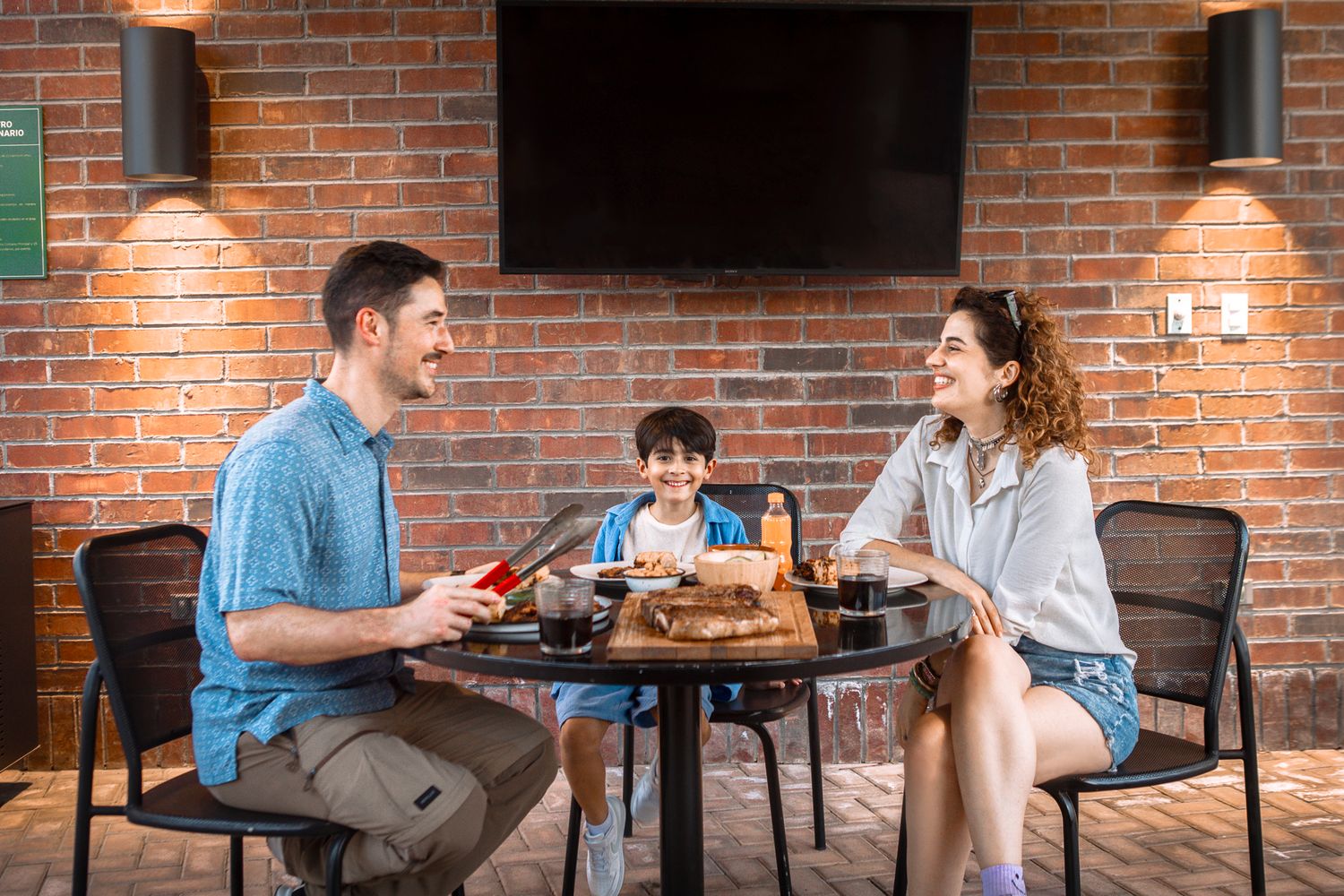 Family dining together at rustic table with exposed brick wall and mounted TV in background.