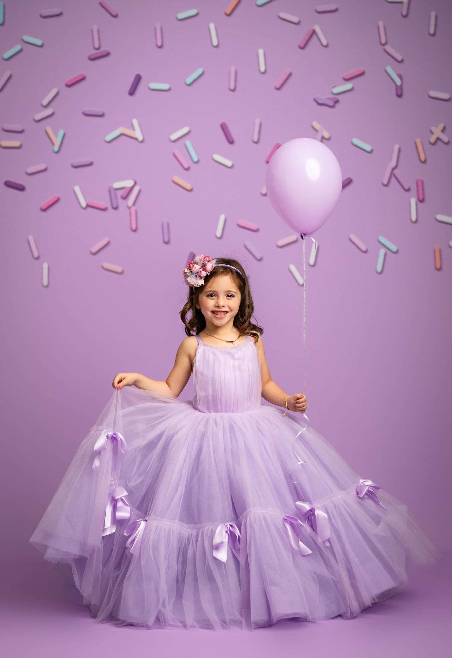 A lavender tulle ball gown with bow accents adorns a young child in a dreamy pastel studio setting.