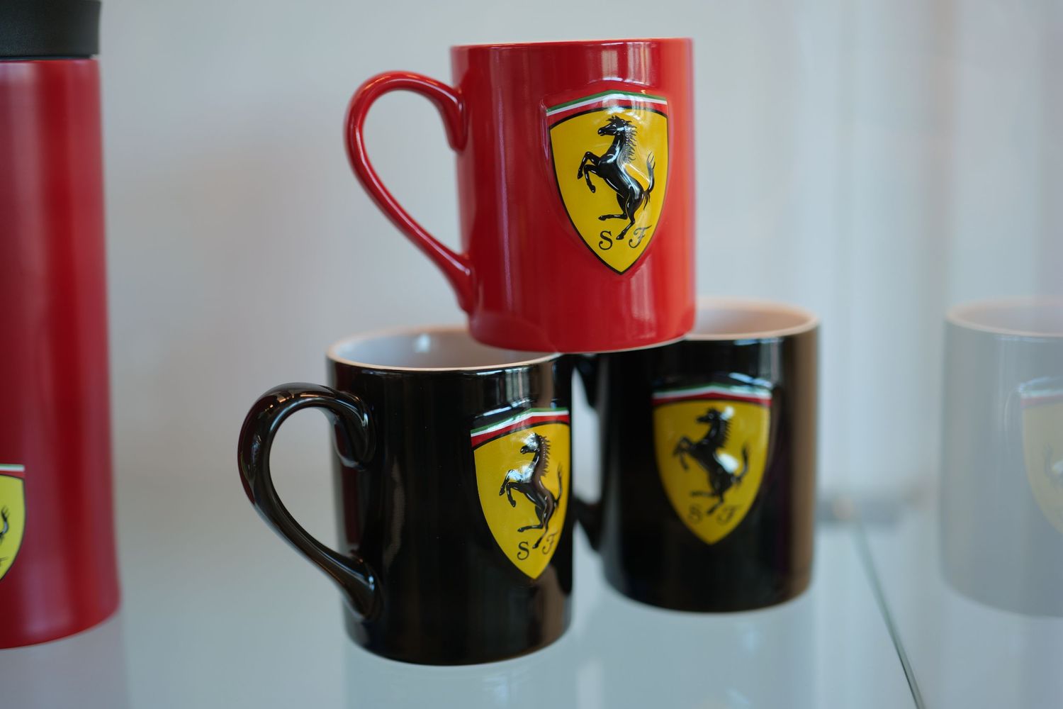 Red and black Ferrari branded ceramic coffee mugs displayed on a glass shelf.