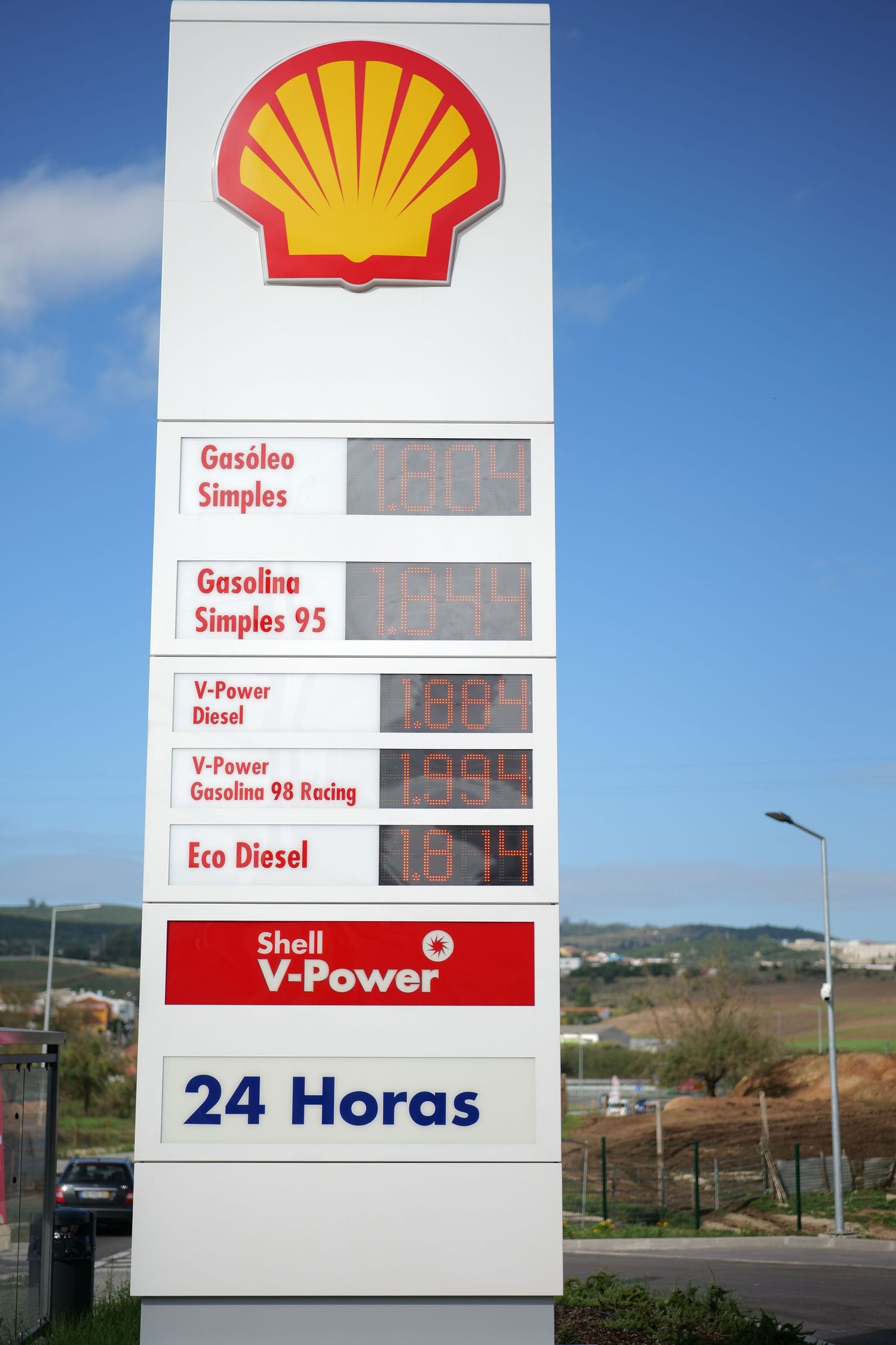 Shell V-Power 24-hour gas station price sign showing fuel prices under blue sky.