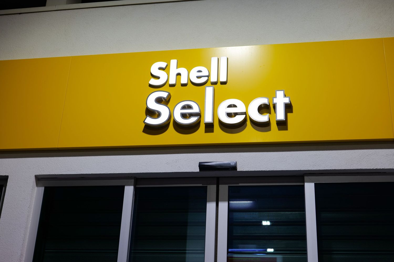 Shell Select convenience store yellow illuminated sign on gas station building facade at night.