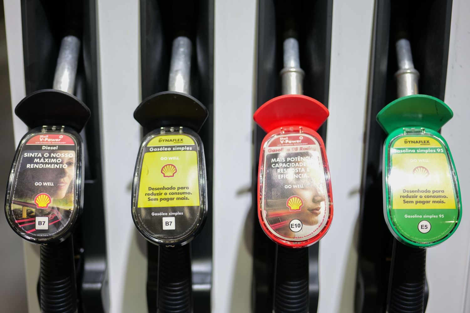 Four colorful Shell fuel pump nozzles in black, yellow, red, and green at a gas station.