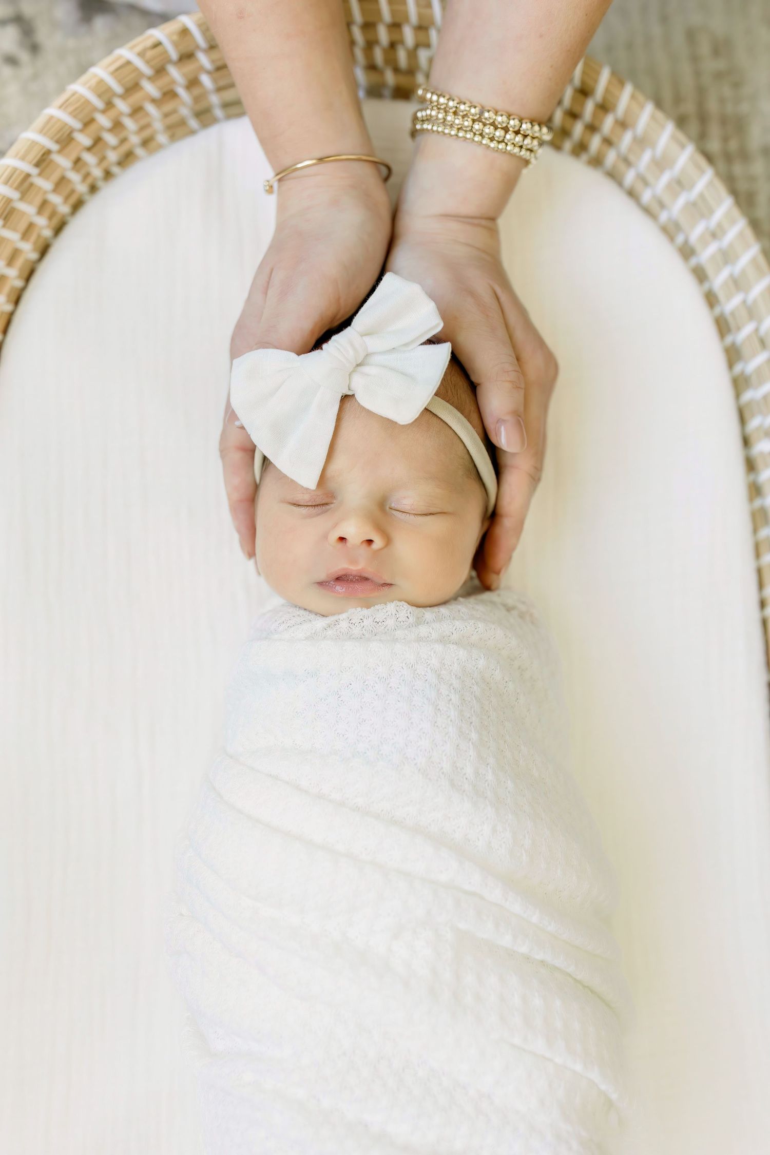 A sleeping newborn swaddled in white with a bow headband resting in a bassinet.