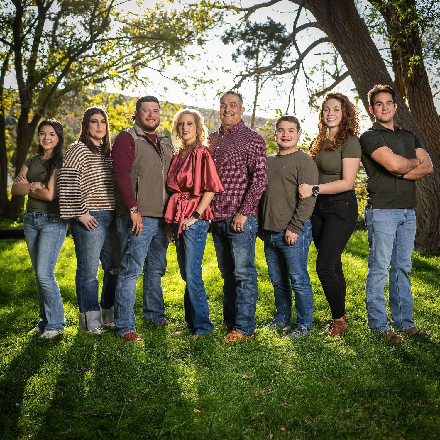 Group of people sitting in a row on grassy hill with trees in background during golden hour lighting.