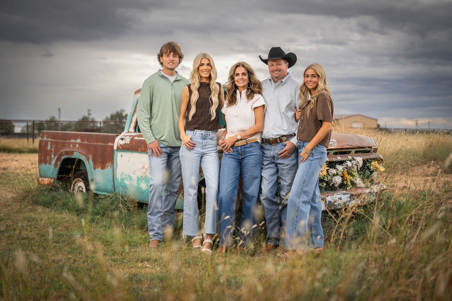 A group stands together in casual western attire against a dramatic cloudy sky in a rural setting.